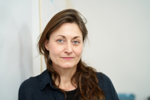 Auf dem Bild ist die Kunsttherapeutin und Systematische Coachin Christa Hammer vor einer weißen Malwand sitzend zu sehen.. Sie blinkt klar und freundlich und hat braune lange Haare und ein schwarzes Oberteil an. Ihr Blick schaut einen direkt an.